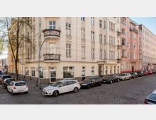 Ecke eines mehrstöckigen Wohnhauses im Altbaustil mit dekorativen Fassadenelementen und Balkonen, umgeben von gepflasterten Straßen. Im Vordergrund parken Autos und Fahrräder; im Erdgeschoss befinden sich große Fenster und ein Geschäftseingang.