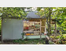 Ein kleines Gartenhaus mit Holzrahmen und großen Glasfenstern, das von Bäumen und Pflanzen umgeben ist. Im Hintergrund sind ein rustikaler Innenraum mit Möbeln sowie eine Terrasse mit Sitzmöbeln sichtbar.