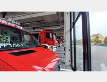 Innenansicht einer Feuerwehrgarage mit mehreren roten Feuerwehrfahrzeugen, die in Reihe geparkt sind. Der Raum hat eine Beton- und Metallkonstruktion mit großen Glasfenstern, die den Blick nach draußen auf eine gepflasterte Straße und Gebäude ermöglichen.