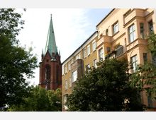 Backstein-Kirchturm mit grünem Spitzdach neben mehreren Wohngebäuden mit hellen Fassaden und Fenstern; im Vordergrund Bäume.