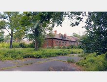 Ein verlassenes Backsteingebäude mit teilweise beschädigtem Dach, umgeben von dichter Vegetation, Bäumen und einem verwilderten Gelände mit Asphaltflächen im Vordergrund.