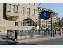 Straßenszene vor einem historischen Gebäude mit heller Steinfassade und verzierter Fensterfront. Im Vordergrund ein U-Bahn-Zugang mit einem Schild „Bhf. Rathaus Neukölln“ und Menschen, die die Straße überqueren oder am Gehweg stehen.