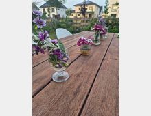 Holz-Gartentisch auf einer Terrasse mit mehreren Glasvasen voller lila- und weißfarbener Blumen; im Hintergrund ein bepflanzter Garten mit Zaun und moderne Wohnhäuser.