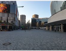 Weitläufiger, gepflasterter Platz in einer modernen Stadtlandschaft, umgeben von Gebäuden mit Glasfassaden, darunter ein Zalando-Gebäude. Links sind Restaurants mit Außensitzplätzen zu sehen, rechts ein Gebäude mit geschwungener Architektur und Eingangsbereich.