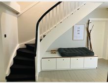 Innenansicht eines Wohnbereichs mit einer weiß gestrichenen Wendeltreppe mit schwarzem Teppichbelag. Darunter befindet sich ein weißer Einbauschrank mit einem gepolsterten Sitzkissen, darüber hängt ein gerahmtes Poster, daneben stehen dekorative Äste an der Wand.