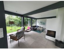 Moderne Wohnzimmergestaltung mit großen, bodentiefen Fenstern, die einen Blick auf einen grünen Garten bieten. Der Raum enthält eine graue Couch, einen weißen Couchtisch, einen Kamin und Sitzmöbel mit Fellauflagen auf einem hellen Fliesenboden.