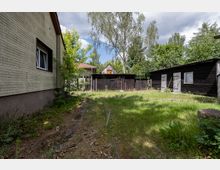Hinterhof mit überwachsenem Rasen und schmalem Weg neben einem hellen Ziegelhaus; im Hintergrund stehen dunkle Holzschuppen mit Toren und Türen. Hohe Bäume und dichtes Grün umgeben das Grundstück unter einem teils bewölkten Himmel.
