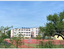 Außenansicht eines Schulgebäudes mit mehreren Etagen und weißen Wänden, umgeben von Bäumen und Sträuchern. Im Vordergrund befindet sich ein Sportplatz mit rotem Kunststoffbelag, der von einem Zaun umgeben ist.