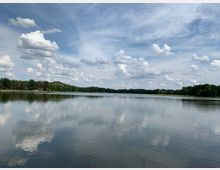Ein ruhiger See mit glatter Wasseroberfläche, die den bewölkten Himmel und umliegende Bäume am Ufer spiegelt. Im Hintergrund dichter Wald unter einem teils bewölkten blauen Himmel.