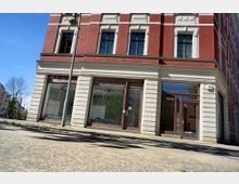 Straßenansicht eines historischen Gebäudes mit roter Ziegelfassade und hoher weißer Sockelverkleidung. Im Erdgeschoss befinden sich große verglaste Schaufenster und eine Eingangstür, daneben eine Hausnummer „22a“.