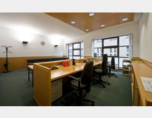 Büro- oder Konferenzraum mit langen Holztischen, mehreren Bürostühlen und einem an der Wand montierten Uhr. Im Hintergrund befinden sich hohe Fenster mit Blick auf ein Backsteingebäude und eine Reihe von Stühlen entlang einer Wand.
