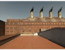 Industriegebäude aus rotem Backstein mit schrägem Dach und markanten Schornsteinen, die mit drehbaren, metallischen Aufsätzen versehen sind. Im Vordergrund ist eine große, mit roten Fliesen ausgelegte Terrasse zu sehen.