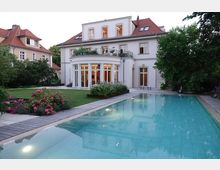 Elegante Villa mit heller Fassade und Satteldach, umgeben von einem gepflegten Garten mit Blumenbeeten. Im Vordergrund ein rechteckiger Swimmingpool, umgeben von einer Terrasse aus Holzplanken.