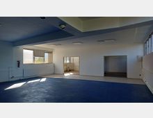 Leerer Innenraum eines Gebäudes mit blauen Bodenbelägen, weißen Wänden und rechteckigen Fenstern, die Tageslicht einlassen. Der Raum verfügt über Heizkörper an den Wänden und offene Durchgänge, die in kleinere angrenzende Räume mit gefliesten Böden führen.