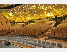 Ein moderner Hörsaal oder Veranstaltungssaal mit ansteigenden Sitzreihen aus Holz und zusätzlichen Stühlen im vorderen Bereich. Die Decke ist mit gelben, wabenförmigen Elementen und zahlreichen kleinen Deckenleuchten gestaltet.