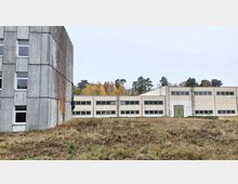 Ein flaches, funktionales Industriegebäude aus Beton, rechts im Bild, mit großen Fenstern und einem grünen Eingangstor. Links ist ein höherer Gebäudeteil mit grauer Fassade zu sehen, leicht bewachsen mit Efeu; im Hintergrund ein Wald mit herbstlich gefärbten Bäumen.