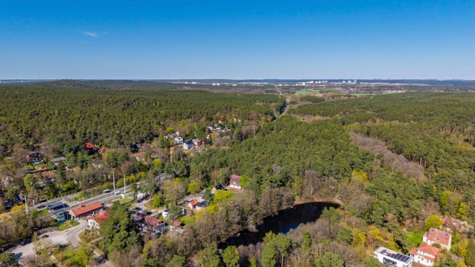 Luftaufnahme einer weitläufigen Waldlandschaft mit verstreuten Wohnhäusern und einer Bahntrasse am Rand einer kleinen Siedlung. Im Vordergrund ist ein dunkler, ovaler Teich von Bäumen umgeben, im Hintergrund reicht der Blick bis zu einer entfernten Stadt am Horizont.