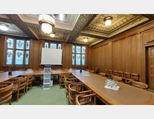 Holzvertäfelter Konferenzraum mit großen, verzierten Fenstern aus Buntglas, einer langen rechteckigen Tischanordnung, Lederstühlen und einer weißen Leinwand auf einem Ständer. Die Decke ist aufwendig gestaltet und mit dekorativen Kronleuchtern beleuchtet.