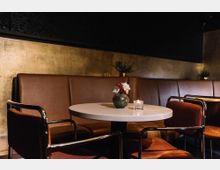 Innenansicht eines schicken Cafés oder Restaurants mit braunen Lederbänken, einem runden weißen Tisch, darauf eine kleine Blumenvase und ein Teelicht, umgeben von Stühlen mit samtartigen, dunkelroten Polstern. Die Wand im Hintergrund hat eine goldfarbene Struktur, dekoriert mit dezenten Pflanzen.