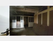 Dunkler Raum in einem scheinbar verlassenen Gebäude mit sichtbaren Deckeninstallationen, Holzwänden und einer Trennwand aus Glas- und Holztüren. Im Hintergrund befindet sich ein weiterer Raum mit Holzwänden.