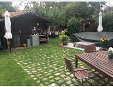 Ein Garten mit Holzhäuschen, umgeben von dichter Vegetation. Im Vordergrund steht eine Sitzgruppe aus Holz auf einem mit Gras durchsetzten Steinboden, daneben eine abgedeckte Fläche, möglicherweise ein Pool.