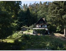 Traditionelles Haus mit Holzfassade und Satteldach inmitten eines Waldes. Daneben steht eine kleine Hütte mit begrüntem Dach, umgeben von Bäumen, Wildwiese und einem offenen Gartenbereich mit Sitzgelegenheiten.