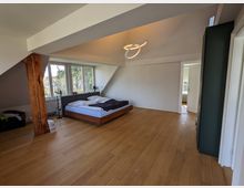 Geräumiges Schlafzimmer im Dachgeschoss mit Holzfußboden, schrägen Deckenflächen und sichtbarem Holzbalken. Ein niedriges Bett steht vor großen Fenstern mit Blick ins Grüne; an der Decke hängt eine moderne Ring-Pendelleuchte, rechts führt eine Tür in einen Nebenraum.