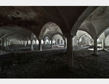 Verlassener Gewölbekeller mit Rundbögen und massiven Säulen, die eine niedrig gewölbte Decke tragen; der Boden ist uneben und von Schutt bedeckt, während Tageslicht durch vereinzelt offene Bereiche einfällt.