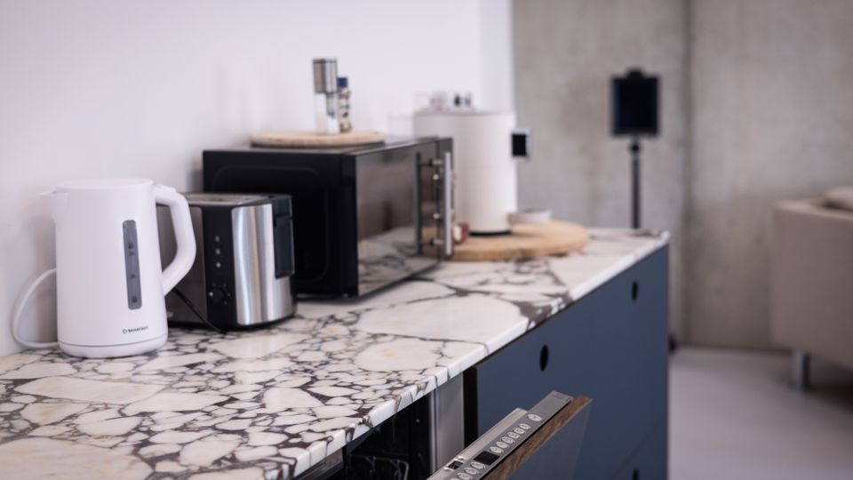Eine moderne Küchenzeile mit einer Arbeitsplatte aus weiß-schwarz gemustertem Marmor, darauf befinden sich ein Wasserkocher, ein Toaster und eine Mikrowelle. Im Hintergrund ist eine graue Betonwand sichtbar, daneben ein Teilbereich eines Sofas.