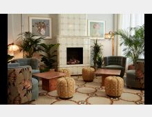 Gemütlicher Lounge-Bereich mit gepolsterten Sesseln, runden Hockern und zwei marmoren Couchtischen. Im Hintergrund steht ein weiß gefliester Kamin mit brennendem Feuer, umgeben von Pflanzen und dekorativen Kunstwerken an der Wand.