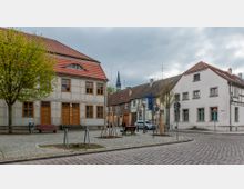 Straßenszene in einer Kleinstadt mit gepflastertem Platz und historischen Gebäuden. Ein Fachwerkhaus mit rotem Ziegeldach und Holzfenstern steht links, daneben weitere Altbauten; im Hintergrund ist ein Kirchturm sichtbar.