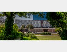 Ein moderner Gebäudekomplex mit geometrischer Fassadengestaltung, bestehend aus weißen und braunen Dreiecksmustern, im Vordergrund von Bäumen und Grünflächen eines Parks umrahmt. Im Hintergrund ist ein hohes Gebäude mit blauer Glasfassade sichtbar.