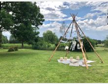 Ein weitläufiger, grüner Garten mit einem Tipi aus Holzstämmen, das mit Lichterketten, weißen Wimpelgirlanden und Traumfängern dekoriert ist. Unter dem Tipi liegen Teppiche und kleine Sitzhocker, umgeben von Bäumen und Büschen unter einem bewölkten Himmel.