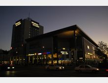 Einkaufszentrum "Spandau Arcaden" bei Dämmerung, mit erleuchteten Schildern verschiedener Geschäfte und Restaurants; ein mehrstöckiges Gebäude mit großem Eingangsbereich und angeschlossenem Turm. Vor dem Gebäude fahren Autos auf einer beleuchteten Straße.