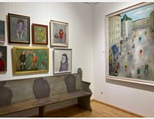Ecke eines Kunstmuseums mit weißen Wänden und hellem Holzboden, an denen mehrere gerahmte Gemälde in Hängung zu sehen sind. Unter den Bildern steht eine lange, bemalte Holzbank, rechts hängt ein großes Gemälde mit einer Straßenszene zwischen hohen Gebäuden.