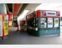 Kleiner Imbissstand mit grünen Kacheln und einem roten Dach in einem überdachten Außenbereich, umgeben von Werbeplakaten und Treppenaufgängen. Imbiss-Schilder und Coca-Cola-Werbung sind prominent sichtbar.