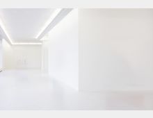 Minimalistischer weißer Raum mit glatten Wänden, hellem Boden und eingebetteten Lichtleisten an der Decke; der Raum wirkt leer und freundlich beleuchtet.