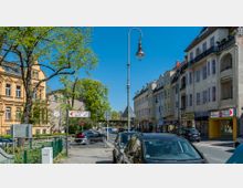 Straßenszene mit mehrstöckigen Gebäuden im historischen Stil links und moderneren Wohn- und Geschäftshäusern rechts, zahlreiche parkende und fahrende Autos, sowie ein Wegweiser zum Museumsdorf Düppel im Vordergrund. Im Hintergrund ist eine Baumallee unter blauem Himmel sichtbar.