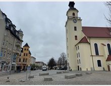 Stadtplatz mit einer großen, hellgelben Kirche mit Turm und Uhr rechts im Bild, umgeben von historischen Wohn- und Geschäftshäusern mit verschiedenen Fassadenstilen. Der Platz ist gepflastert und von einzelnen Betonbänken sowie parkenden Fahrzeugen gesäumt.