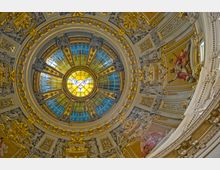 Deckenansicht einer reich verzierten Kuppel aus einer Kirche, mit einem zentralen Buntglasfenster, das eine Taube symbolisiert, umgeben von goldenen Details, Mosaiken und skulpturalen Ornamenten. Die blauen und gelben Glasabschnitte kontrastieren mit den goldenen und cremefarbenen Verzierungen.