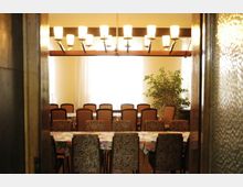 Ein Speiseraum mit mehreren gleichmäßig angeordneten Tischreihen, die mit geblümten Tischdecken bedeckt sind, und Polsterstühlen. Im Hintergrund sind große Fenster mit transparenten Vorhängen, eine hängende Deckenleuchte mit mehreren Lampen und eine große Zimmerpflanze zu sehen.