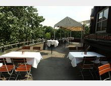 Dachterrasse eines Restaurants mit mehreren Tischen mit weißen Tischdecken und Holzklappstühlen, teilweise unter einem weißen Pavillonzelt. Links grenzt ein Geländer an dichtes Grün, rechts verläuft die Fassade des Gebäudes mit Fenstern.