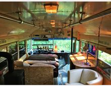 Innenraum eines umgebauten Schulbusses mit Wohnbereich. Zu sehen sind ein Bett, eine kleine Sitzecke mit weißem Sessel, ein Tisch mit Lampe und die Fahrerkabine im Vordergrund; durch die Fenster ist eine grüne Landschaft zu erkennen.