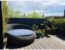 Eine Terrasse mit Holzplankenboden, auf der sich ein großer, runder Rattan-Loungesessel mit blauem Sitzkissen befindet. Umgeben von verschiedenen Pflanzen in Töpfen und Kästen, im Hintergrund eine niedrige Brüstungsmauer und Bäume unter einem bewölkten Himmel.