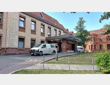 Eingangsbereich einer Klinik mit der Beschilderung "Notaufnahme", links ein modernes Gebäude mit beigefarbener Fassade und rechts ein historisches Backsteingebäude. Vor dem Gebäude stehen ein Krankenwagen und ein Transporter auf einer gepflasterten Zufahrt, umgeben von Bäumen und Grünflächen.