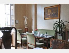 Innenraum eines eleganten Cafés oder Salons mit klassischen Möbeln. Ein Marmortisch ist von grünen Polsterstühlen umgeben, an den beige-gemusterten Wänden befinden sich ein Gemälde in einem goldenen Rahmen und eine Statue auf einem Sockel, daneben eine Zimmerpflanze auf einem Podest.