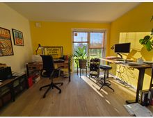 Helles Arbeitszimmer mit gelb gestrichener Wand, einem höhenverstellbaren Schreibtisch, Bildschirm und Bürostuhl. Große Fenster im Hintergrund mit Blick ins Freie, Zimmerpflanzen und Wanddekoration ergänzen die Einrichtung.