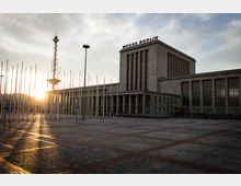 Außenansicht des Messegebäudes in Berlin mit der Aufschrift "Messe Berlin" auf dem Dach, im Vordergrund ein gepflasterter Platz mit leeren Fahnenmasten. Im Hintergrund ist der Funkturm sichtbar, während die tiefstehende Sonne durch die Masten scheint.