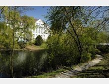 Großes, weißes Gebäude mit klassizistischer Architektur, darunter hohe Fenster und ein Satteldach, im Hintergrund eines Parks mit einem Flussufer. Davor ein schmaler, gepflasterter Weg umgeben von Bäumen und Grünflächen.
