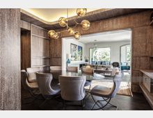 Modernes Esszimmer mit ovalem Glastisch und gepolsterten Stühlen, umgeben von holzverkleideten Wänden. Im Hintergrund ist ein helles Wohnzimmer mit großen Fenstern, einer Sofagruppe und dekorativen Elementen zu sehen.